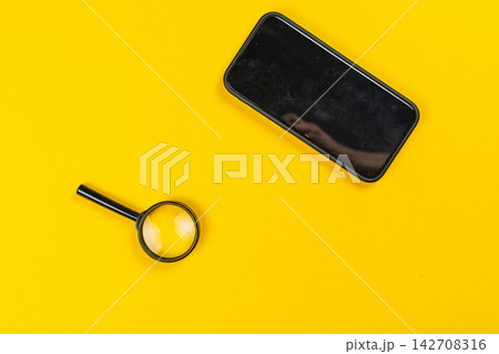 Using a magnifying glass to examine a smartphone on a yellow background 142708316