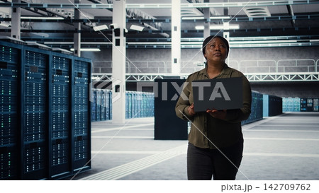 Data center engineer using laptop to develop disaster recovery plans, minimizing downtime. Server farm IT expert enhancing data protection protocols using notebook, camera A 142709762