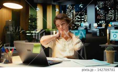 Businesswoman stretching her upper back and neck muscles, correcting the posture and neck stiffness for relief and relaxation. Intense office activity due to overtime late at night. Camera A. Businesswoman stretching her upper back and neck muscles, correcting the posture and neck stiffness for relief and relaxation. Intense office activity due to overtime late at night. Camera A. 142709781