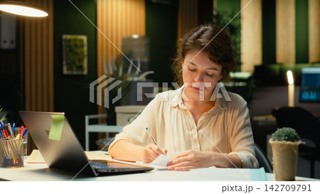 Livid businesswoman breaking a paper page after making a mistake, handwriting and notetaking in office after hours. Displeased impatient worker redoing the draft on a new sheet. Camera B. Livid businesswoman breaking a paper page after making a mistake, handwriting and notetaking in office after hours. Displeased impatient worker redoing the draft on a new sheet. Camera B. 142709791