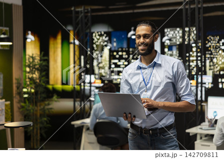 Smiling Arab employee standing in office, reading emails on laptop screen, responding to inbox messages. Happy man analyzing data on notebook display at night, interpreting statistical information 142709811