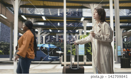Woman traveler holding takeaway coffee pulls luggage while walking inside bus terminal. Tourist enjoying refreshment before continuing intercity journey with shuttle 142709841