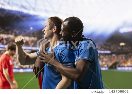 Happy diverse football players celebrating winning goal on pitch, with arms raised and clenched fists. Devastated opponents while rivals hugging teammates and celebrating with cheering fans. 142709901