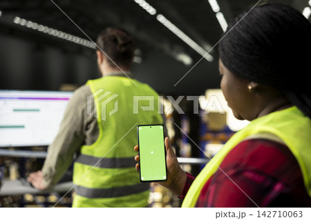 Vertical green screen phone held by female staff involved in logistics and order fulfillment, surrounded by organized shelves. Supply chain management with shipping operations and safety practices. 142710063