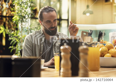 Freelancer man using laptop at kitchen table and working remotely, casual professional lifestyle with online productivity and digital multitasking in a rustic home setup with natural light. Freelancer man using laptop at kitchen table and working remotely, casual professional lifestyle with online productivity and digital multitasking in a rustic home setup with natural light. 142710064