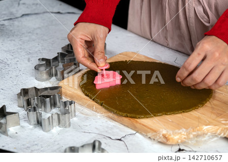 Hands cutting rolled dough with pink cookie cutter on wooden board, surrounded by metal cutters for baking, pastry preparation, homemade cookies and festive kitchen cooking Hands cutting rolled dough with pink cookie cutter on wooden board, surrounded by metal cutters for baking, pastry preparation, homemade cookies and festive kitchen cooking 142710657