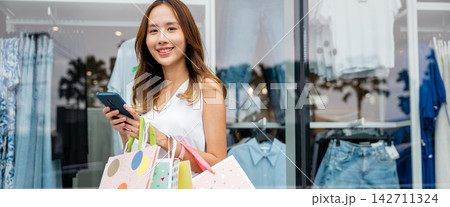 woman holding shopping bags and a cellphone smiles in front of a store window 142711324
