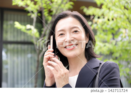 屋外でスマホを使って電話する50代のビジネスウーマン、外出先で仕事の連絡をする働く女性クローズアップ 142711724