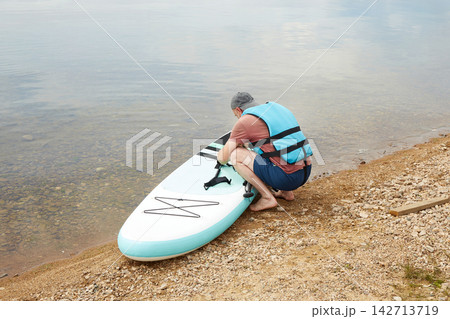 A man in a life jacket is getting ready to paddleboard on the edge of a lake. A man in a life jacket is getting ready to paddleboard on the edge of a lake. 142713719
