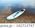 Inflatable stand up paddleboard (SUP board) with paddle resting on a rocky lakeshore, partly in clear calm water. 142713747