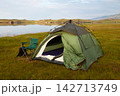 A green tent is set up on a grassy area by a calm lake. A chair is positioned next to the tent.  142713749