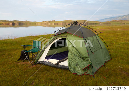 A green tent is set up on a grassy area by a calm lake. A chair is positioned next to the tent. A green tent is set up on a grassy area by a calm lake. A chair is positioned next to the tent. 142713749