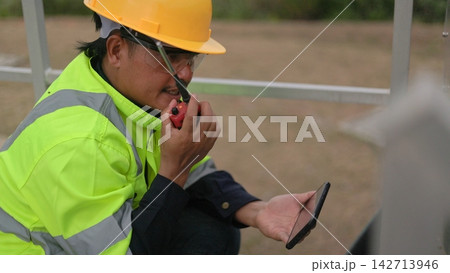 Close-up shot of an Asian male engineer wearing safety hardhat and vest using a walkie talkie radio and checking a smartphone at an outdoor industrial energy power plant. 142713946