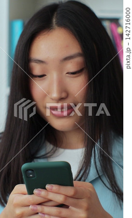 Asian woman took a break between studies and using smartphone, texting message while standing in the library. University library concept. Asian woman took a break between studies and using smartphone, texting message while standing in the library. University library concept. 142716000