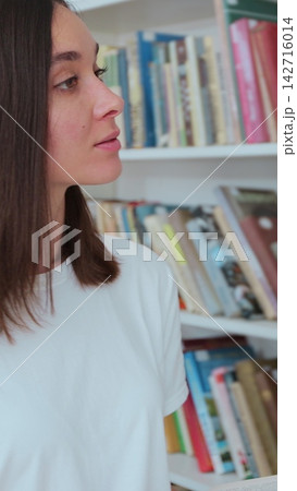 Caucasian girl in white t-shirt walks along the shelves with books in the library. University library concept. Caucasian girl in white t-shirt walks along the shelves with books in the library. University library concept. 142716014