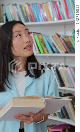 Asian girl in blue shirt walks along the shelves with books in the library. College education concept. Asian girl in blue shirt walks along the shelves with books in the library. College education concept. 142716015