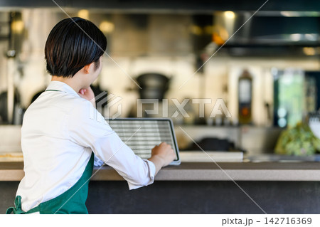 タブレットを見る店員、カウンター越しに厨房を望む タブレットを見る店員、カウンター越しに厨房を望む 142716369