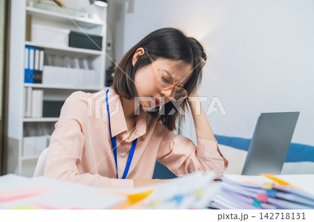 Stressed young Asian office worker overwhelmed by paperwork while working on a laptop. Stressed young Asian office worker overwhelmed by paperwork while working on a laptop. 142718131