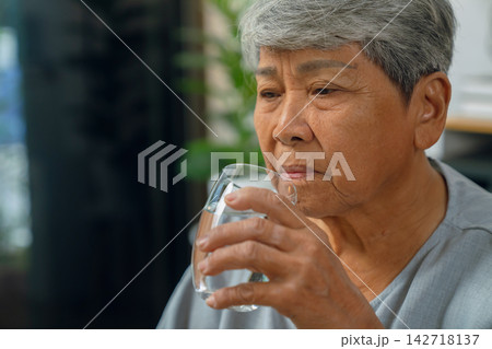 Elderly Asian woman drinks water on a sofa at home, promoting hydration and healthy living. Elderly Asian woman drinks water on a sofa at home, promoting hydration and healthy living. 142718137