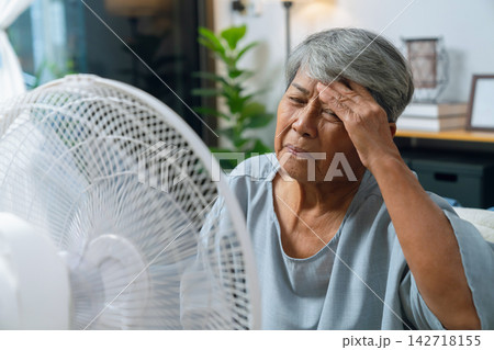 Elderly Asian woman cools off with a fan in her overheated living room. Elderly Asian woman cools off with a fan in her overheated living room. 142718155