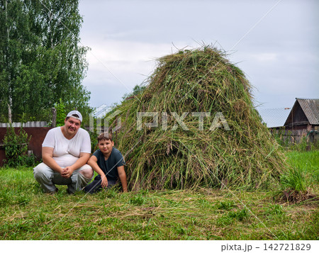 Father and son crouched down in the grass next to a large, freshly built haystack, celebrating a successful harvest on a rural farm during a cloudy summer day. Father and son crouched down in the grass next to a large, freshly built haystack, celebrating a successful harvest on a rural farm during a cloudy summer day. 142721829