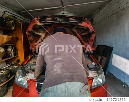 Man in work gloves and dirty clothes leaning over a car with its hood open, checking the engine during an auto service in a messy private garage. Man in work gloves and dirty clothes leaning over a car with its hood open, checking the engine during an auto service in a messy private garage. 142721833