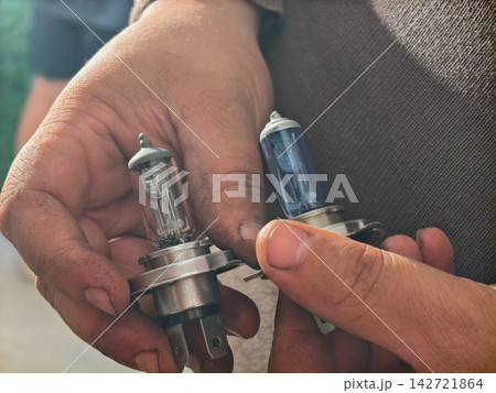 Mechanic hands holding and comparing two automotive halogen headlight bulbs, a clear old one and a new blue-tinted one, demonstrating car maintenance and replacement. Mechanic hands holding and comparing two automotive halogen headlight bulbs, a clear old one and a new blue-tinted one, demonstrating car maintenance and replacement. 142721864