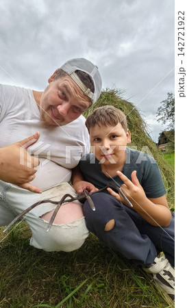 Smiling father and boy sitting next to a pile of fresh hay, holding a traditional pitchfork, enjoying farm work together under a cloudy summer sky. 142721922