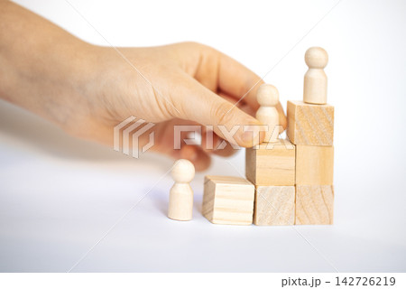 Hand placing wooden figurine on top of stacked blocks, depicting a small staircase made of cubes and cylinders, with additional figurines at the base 142726219