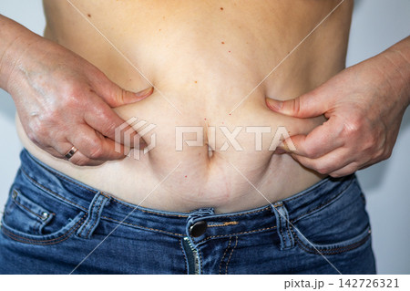 Middle-aged woman pinching belly fat above jeans showing abdominal skin and concern about weight 142726321