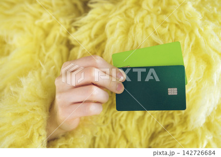 Hand holding two credit cards, one green and one dark blue, against a background of fluffy yellow fabric, emphasizing modern financial transactions and consumerism Hand holding two credit cards, one green and one dark blue, against a background of fluffy yellow fabric, emphasizing modern financial transactions and consumerism 142726684