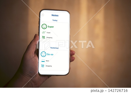 Smartphone displaying notes application with daily tasks and mood indicators held in hand against a blurred background, emphasizing personal organization and productivity 142726716