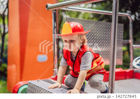 Little boy dressed as firefighter playing in a red toy fire truck at an outdoor playground with helmet safety vest pretend rescue game childhood dreams role play and wide view. Little boy dressed as firefighter playing in a red toy fire truck at an outdoor playground with helmet safety vest pretend rescue game childhood dreams role play and wide view. 142727645