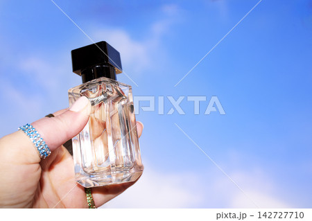 Hand holding a clear glass perfume bottle with a black cap against a bright blue sky, showcasing the elegant design and transparency of the fragrance container 142727710