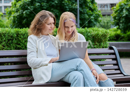 Mother and daughter using laptop together sitting on bench in summer park Mother and daughter using laptop together sitting on bench in summer park 142728039