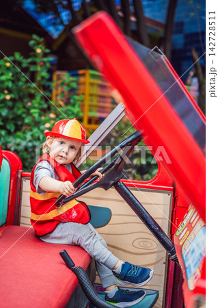 Little boy dressed as firefighter playing in a red toy fire truck at an outdoor playground with helmet safety vest pretend rescue game childhood dreams role play and playing actively. Little boy dressed as firefighter playing in a red toy fire truck at an outdoor playground with helmet safety vest pretend rescue game childhood dreams role play and playing actively. 142728511