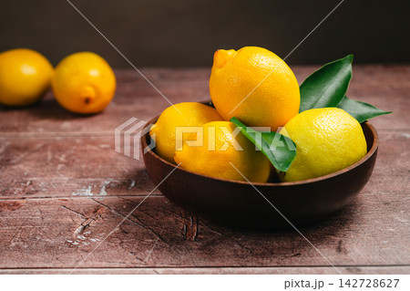 Whole ripe lemons with fresh green leaves in wooden bowl, natural citrus fruit on rustic table 142728627