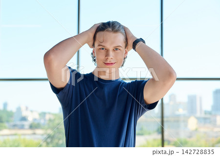 Handsome young man touching hair looking at camera near big window with city view Handsome young man touching hair looking at camera near big window with city view 142728835