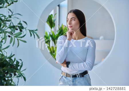 Thoughtful young woman with hand on chin looking away in modern interior 142728881