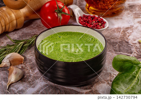 Green spinach soup in black ceramic bowl surrounded by fresh ingredients including tomato, garlic, rosemary, and pink peppercorns on marble surface 142730794