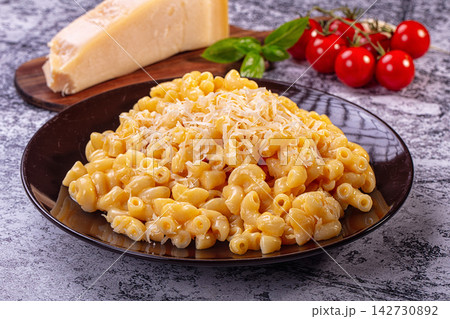 Creamy mac and cheese served in dark bowl with grated parmesan, cherry tomatoes and basil on wooden board against textured gray background Creamy mac and cheese served in dark bowl with grated parmesan, cherry tomatoes and basil on wooden board against textured gray background 142730892