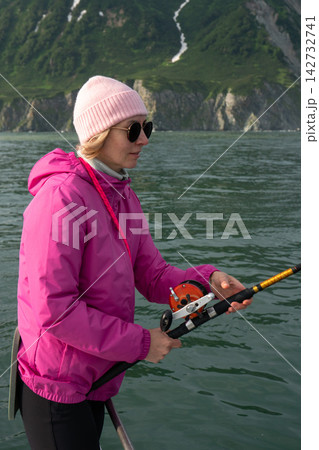 Woman fishes from a boat off Kamchatka coast with green mountains behind. Fishing, sea trip, active travel, outdoor recreation, adventure tourism and memorable journey in remote nature. Woman fishes from a boat off Kamchatka coast with green mountains behind. Fishing, sea trip, active travel, outdoor recreation, adventure tourism and memorable journey in remote nature. 142732741