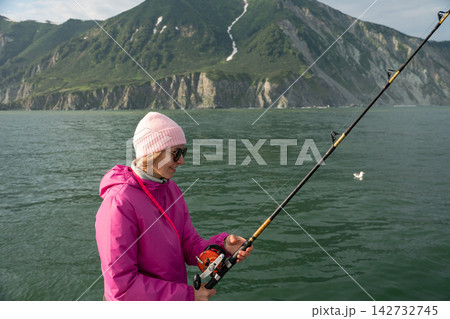 Female traveler fishes from a boat during a scenic sea journey in Kamchatka with rugged green coast behind. Fishing adventure, active tourism, outdoor leisure, exploration and nature travel. Female traveler fishes from a boat during a scenic sea journey in Kamchatka with rugged green coast behind. Fishing adventure, active tourism, outdoor leisure, exploration and nature travel. 142732745