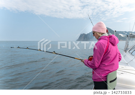 Female angler fishes from a boat beside rugged Kamchatka coast. Scenic sea trip, marine travel, recreational fishing, outdoor leisure, active journey, nature tourism and adventure travel. Female angler fishes from a boat beside rugged Kamchatka coast. Scenic sea trip, marine travel, recreational fishing, outdoor leisure, active journey, nature tourism and adventure travel. 142733833