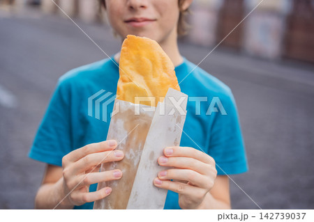 Boy holding a golden molote poblano traditional fried Mexican empanada outdoors on a street in Puebla Mexico with colonial city background paper wrapper local street food authentic travel snack. Boy holding a golden molote poblano traditional fried Mexican empanada outdoors on a street in Puebla Mexico with colonial city background paper wrapper local street food authentic travel snack. 142739037