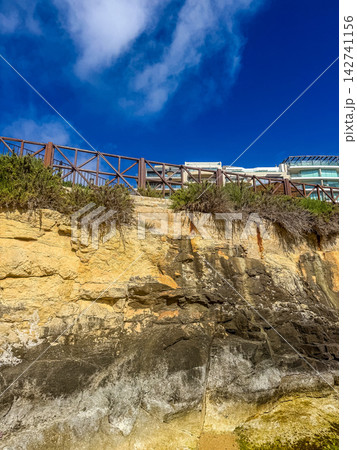 Dramatic rocky cliff face with wooden safety fence, modern hotel building above, blue sky with clouds, coastal Mediterranean landscape. High quality photo Dramatic rocky cliff face with wooden safety fence, modern hotel building above, blue sky with clouds, coastal Mediterranean landscape. High quality photo 142741156