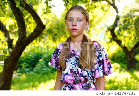 Portrait of a young blonde teen girl in a colorful floral dress standing outdoors in a sunny green garden. Childhood and summer concept Portrait of a young blonde teen girl in a colorful floral dress standing outdoors in a sunny green garden. Childhood and summer concept 142747138