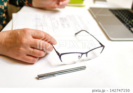 Close-up of a person hands holding glasses on a desk with documents and a laptop. Office work, business, and paperwork concept Close-up of a person hands holding glasses on a desk with documents and a laptop. Office work, business, and paperwork concept 142747150