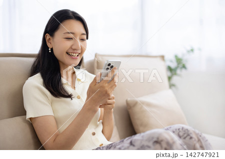 ソファに座りながら笑顔でスマートフォンを操作する若い日本人女性 142747921