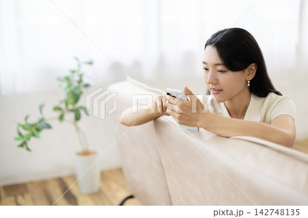 明るい窓辺のソファでスマートフォンを操作する若い日本人女性 明るい窓辺のソファでスマートフォンを操作する若い日本人女性 142748135